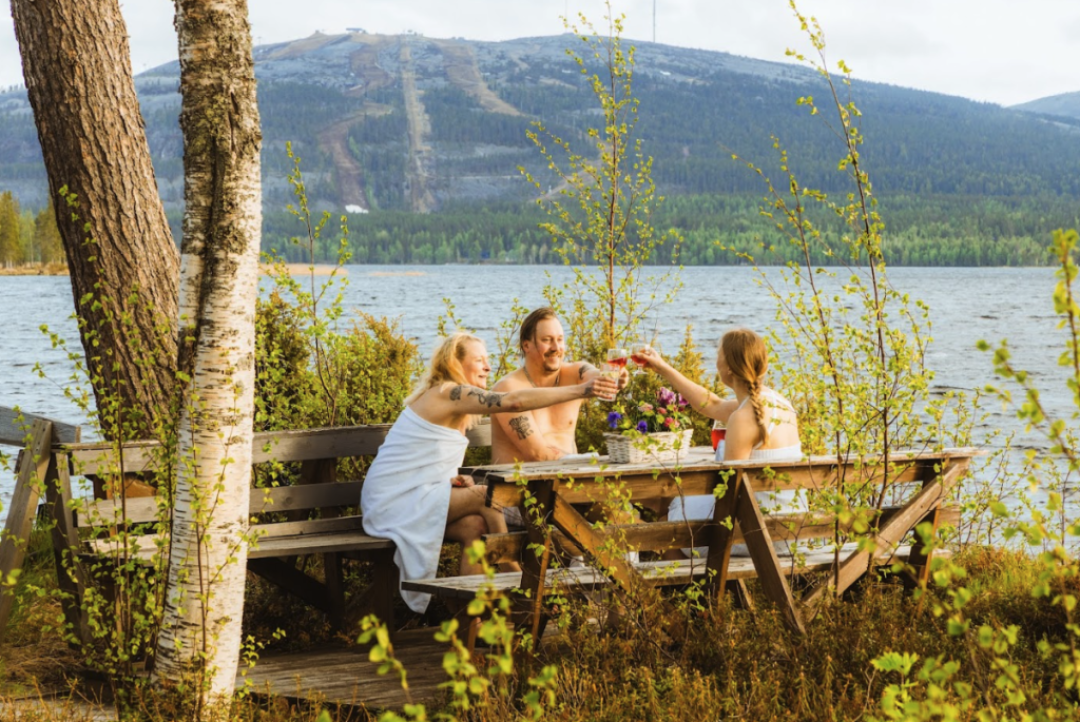 Kolme henkilöä kilistelemässä kesäisen Pyhäjärven rannalla saunan lomassa, Pyhätunturi siintäen horisontissa. 