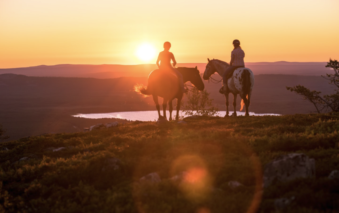 Two people riding horses western style lit by the Midnight Sun of Lappish summer, looking down to the lake Pyhäjärvi from top of a fell.