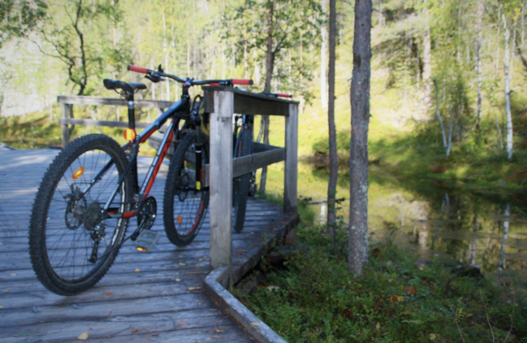 Maastopyörä nojailemassa puisen rakenteen aitaan joen/puron varrella loppu kesän vihreässä maisemassa