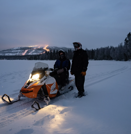 Snowmobile parked on the ice of Pyhäjärvi with a driver and a guide standing next to it, with lights of Pyhä ski slopes in the horizon