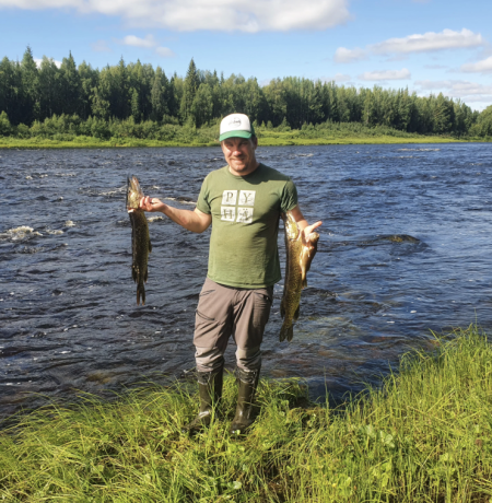 Happy man holding two pikes that he just catched from the raging river behind him with summer greenery under his feet and in the forest behind the river, blue sky above 