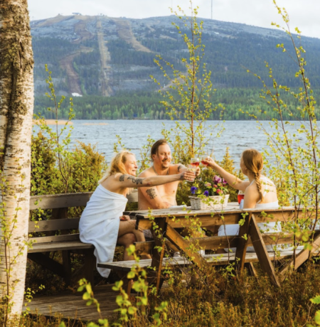 Kolme henkilöä kilistelemässä kesäisen Pyhäjärven rannalla saunan lomassa, Pyhätunturi siintäen horisontissa. 