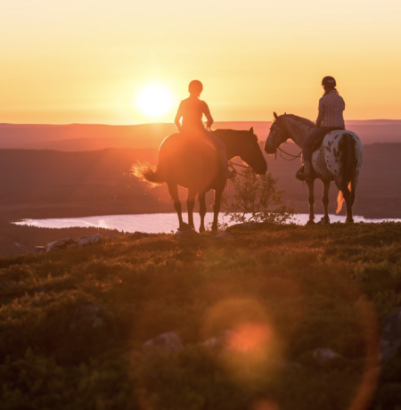 Two people riding horses western style lit by the Midnight Sun of Lappish summer, looking down to the lake Pyhäjärvi from top of a fell.