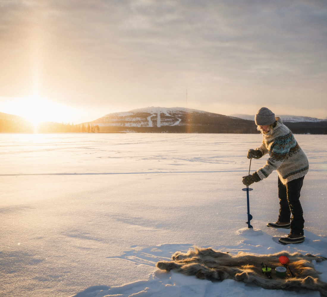A person drilling a hole to the ice of Pyhäjärvi with a bright sunlight shining through the forest in the back