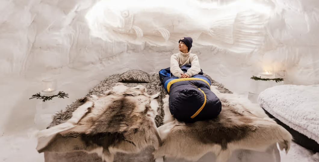 A person is laying in a bed made of ice, on a reindeer fur inside a snow igloo