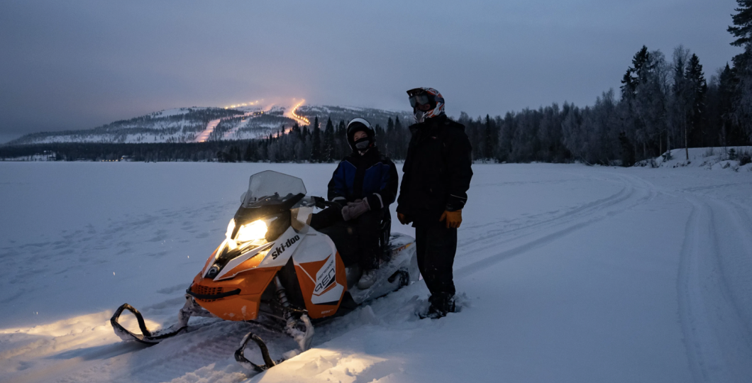 Snowmobile parked on the ice of Pyhäjärvi with a driver and a guide standing next to it, with lights of Pyhä ski slopes in the horizon