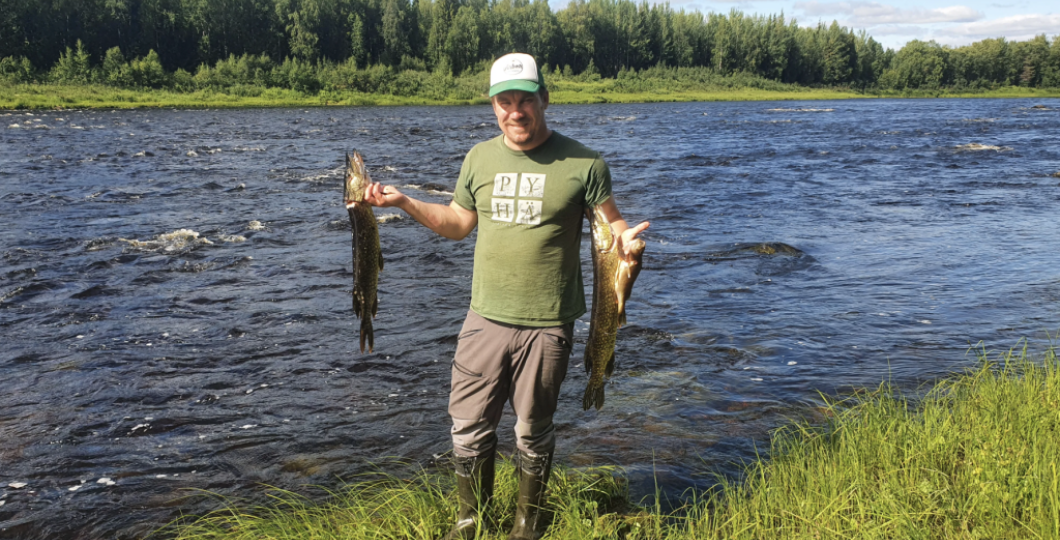 Happy man holding two pikes that he just catched from the raging river behind him with summer greenery under his feet and in the forest behind the river, blue sky above 