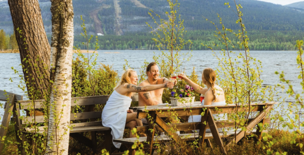 Kolme henkilöä kilistelemässä kesäisen Pyhäjärven rannalla saunan lomassa, Pyhätunturi siintäen horisontissa. 