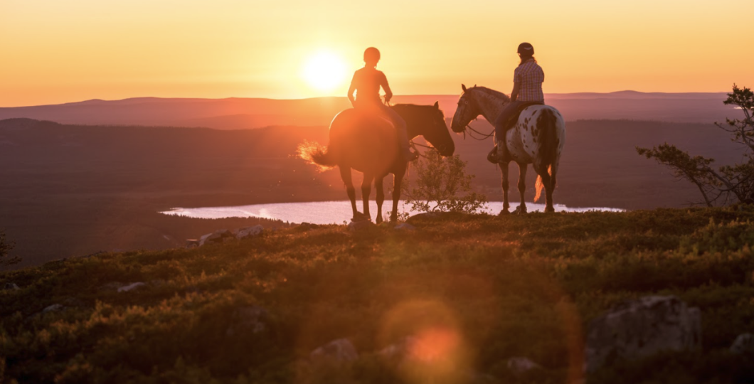Two people riding horses western style lit by the Midnight Sun of Lappish summer, looking down to the lake Pyhäjärvi from top of a fell.