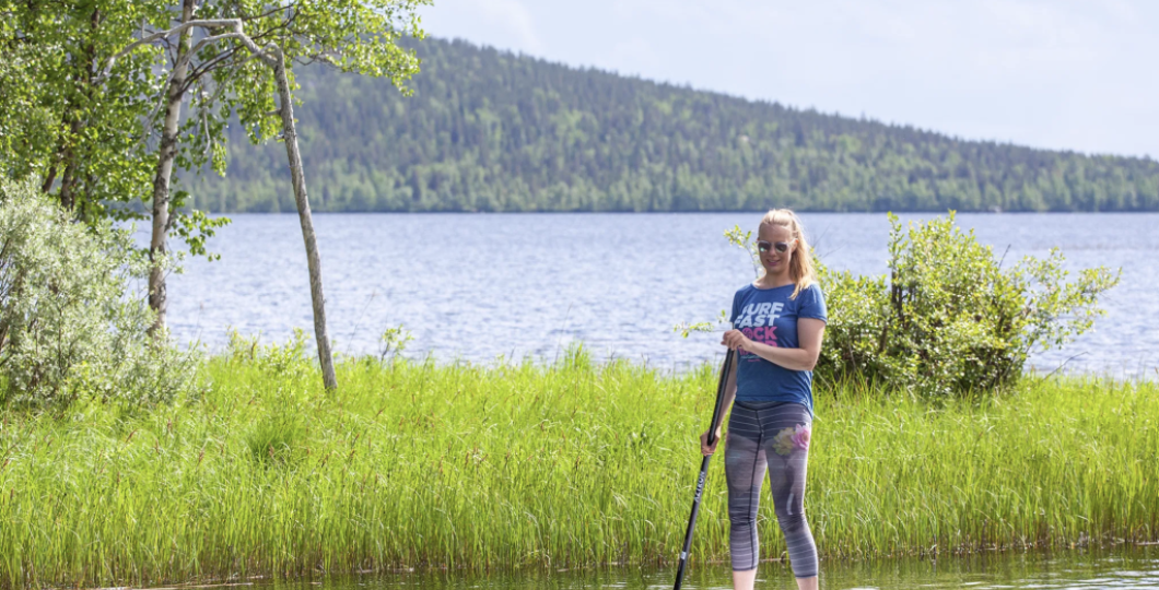 Yksi henkilö seisoo SUP-laudan päällä meloen rauhallisesti Pyhäjärveltä Pyhäjoelle kesäisellä puolipilvisellä ja lähes täysin tyynellä säällä, vihreä kaislikko ja koivun oksat ympäröiden häntä