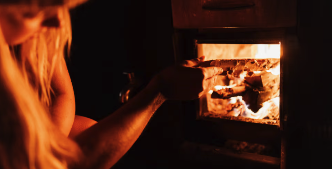 Henkilö laittamassa saunan loimuavaan kiukaan tulipesään lisää puita. 